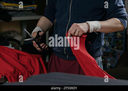 photo of a man with scissors cutting off a piece of red cloth. serigraphy production. printing images on t-shirts by silkscreen method in a design stu Stock Photo