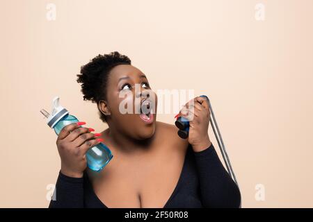 shocked african american oversized woman in sportswear holding sports bottle with water and skipping rope isolated on beige Stock Photo