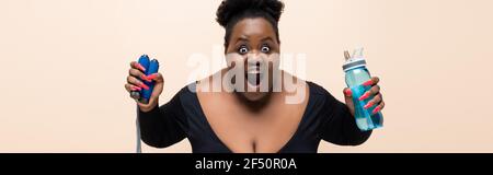 shocked african american plus size woman holding sports bottle and skipping rope isolated on beige, banner Stock Photo