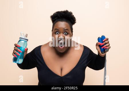 shocked african american plus size woman in sportswear holding sports bottle and skipping rope isolated on beige Stock Photo