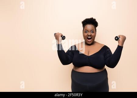 amazed african american plus size woman in sportswear holding dumbbells isolated on beige Stock Photo