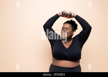 happy african american plus size woman in sportswear holding dumbbells above head isolated on beige