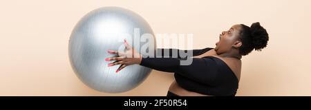 surprised african american plus size woman in sportswear holding fitness ball isolated on beige, banner Stock Photo