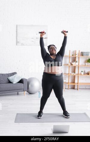 full length of african american plus size woman in sportswear exercising with dumbbells in living room Stock Photo