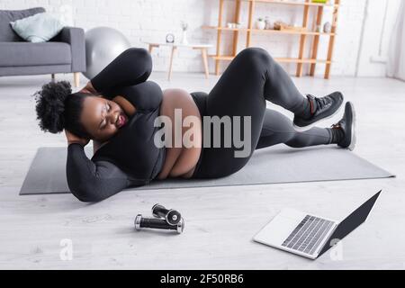 happy african american plus size woman in sportswear working out on fitness mat and looking at laptop