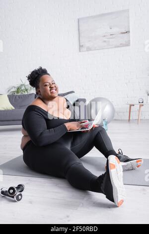 happy african american plus size woman in sportswear holding laptop and sitting on fitness mat Stock Photo