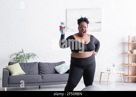 amazed african american plus size woman in sportswear holding sports bottle at home Stock Photo