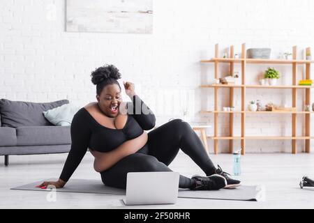 excited african american plus size woman in sportswear sitting on fitness mat and looking at laptop in living room Stock Photo