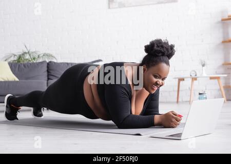 happy african american plus size woman watching online sport training on laptop while doing plank in living room Stock Photo