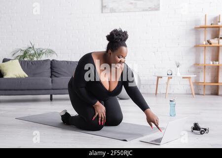 happy african american plus size woman watching sport training on laptop while sitting on fitness mat in living room Stock Photo
