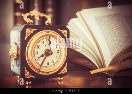 Old wooden alarm clock with gold applications, showing 8 o'clock on a wooden table with books in the background Stock Photo