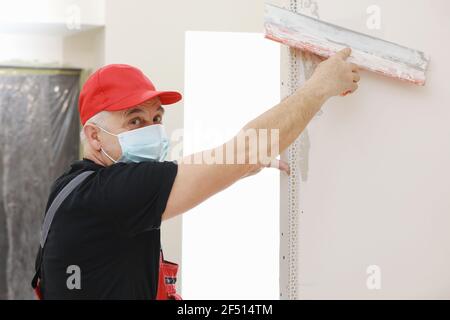Builder in protective mask using a trowel to add plaster. Plastering wall with putty-knife, close up image. Fixing wall surface and preparation for pa Stock Photo