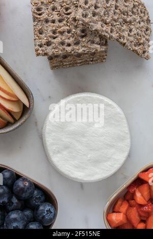 A rounded fresh cheese surrounded by a pile of healthy crackers and three trays full of fruit. Stock Photo