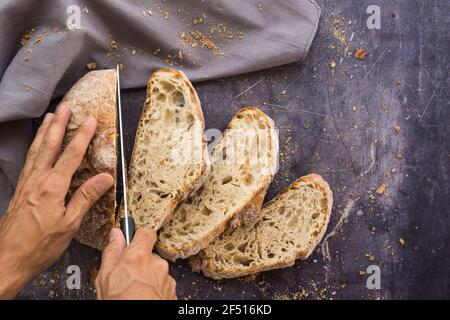 Zenithal image of an organic bread with hands with a knife breaking it into slices placed in a fan shape on a dark surface surrounded by crumbs and a Stock Photo