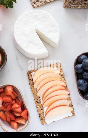 Overhead view of an almost whole fresh cheese with a healthy cracker sheet in the middle with some apple slices on it next to some trays full of fruit Stock Photo