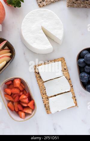 Overhead view of an almost whole fresh cheese with a healthy cracker sheet in the middle with some cheese slices on it next to some trays full of frui Stock Photo