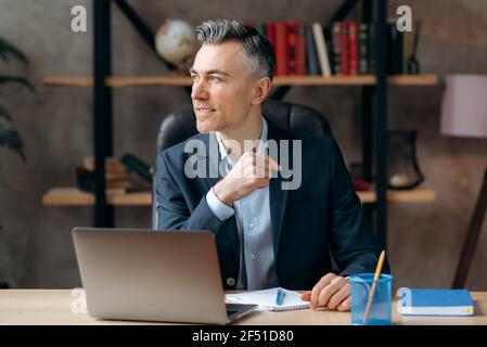 Attractive successful self-confident caucasian adult business leader, agent or lawyer, sitting in modern office, looking out the window, thinking about a business project or dreaming about vacation Stock Photo