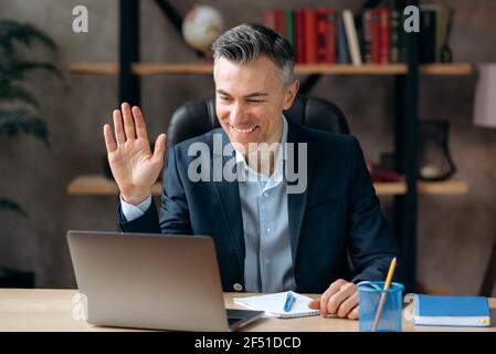 Gray-haired successful pleasant caucasian mature ceo, lawyer or top manager, in formal suit, communicates via video call, conducts online briefing with employees, greeting, smiles friendly Stock Photo