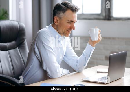 A respectable confident caucasian man, businessman, gray-haired male boss, working on laptop, browsing internet, holding a cup of coffee in his hand, take a pause from work, smiling Stock Photo