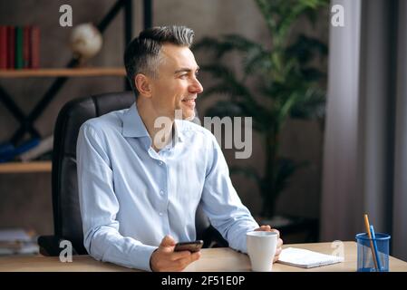 Satisfied successful caucasian senior business leader or manager in formal shirt, took a break during work, sits at the office, holds a smartphone in hand, thinking, pensively looking away, smiling Stock Photo