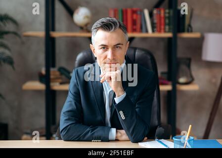 Portrait of a caucasian influential successful gray-haired ceo, lawyer or business leader, focused looks at the camera, sitting at the office, dressed in a formal suit, pleasant smiles Stock Photo
