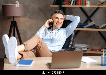 Attractive successful gray-haired businessman, freelancer or manager is talking the phone, calling friends or colleagues, throwing his feet on the table, relaxing office, looks away, smiling happily Stock Photo