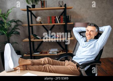 Successful handsome mature caucasian businessman or manager, freelance relaxes while sitting with his legs thrown on his desk, looking at the camera, with hands behind his head, smiling friendly Stock Photo