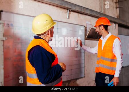 Young engineer and elderly worker in orange vests and hard hat make production plans Stock Photo