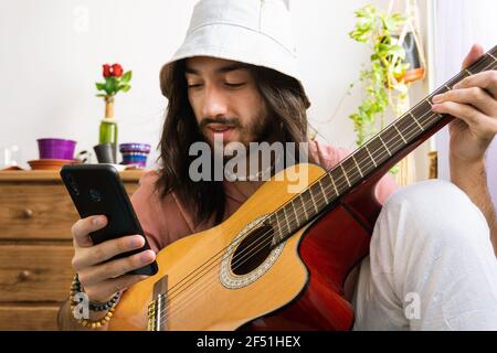 Hipster Guitarist Man Using His Phone Sitting On The Couch At Home. Stock Photo
