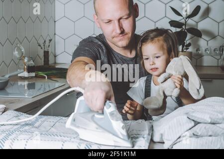 Man ironing bed linen with little daughter on his knees. Father engaged in household chores. Stock Photo