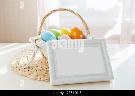 Easter colorful eggs in busket with empty frame on white wooden table. Happy Easter background. Copyspace for text Stock Photo