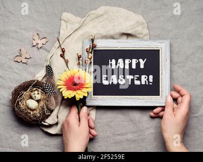 Happy Easter, chalk text on black board. Hands holding blackboard and orange gerbera flower. Quail eggs in bird nest, spring decor. Flat lay, linen Stock Photo