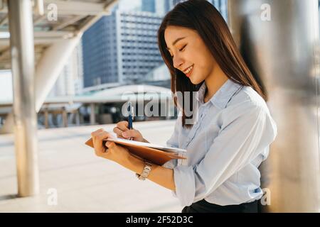 Cheerful young Asian female designer doodling creativity and designs in book while relaxing outside office building during break hours Stock Photo