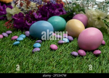 Pastel colored Easter eggs and pastel colored chocolate candy on green grass , against colored flowers background. Stock Photo