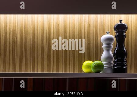 Salt and pepper grinders in black and white on a kitchen shelf with a lime and a lemon fruit on a background of striped brown wallpaper. Stock Photo