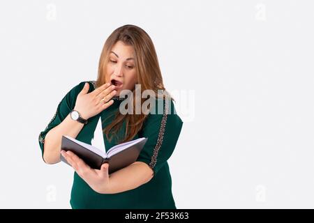 Surprised young overweight woman, gesturing surprise, reading a exciting book while sitting at the library, grey background with side space for Stock Photo