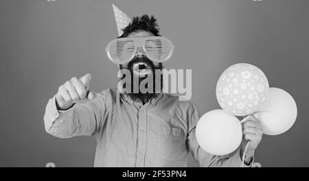 Hipster in giant sunglasses celebrating birthday. Man with beard and mustache on happy face holds bunch of air balloons, red background. Guy in party Stock Photo