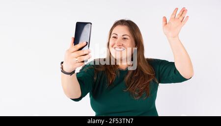 Happy full sized woman smiles and takes a selfie on her smartphone waving and posing. Studio white background. Banner. Stock Photo