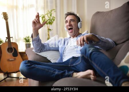 Euphoric adult man laying dancing in sofa listening to music from a mobile with white headphones dressed casual in the living room with window and bac Stock Photo