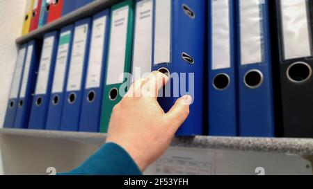 Man hand reaching for a blue office Paper folder in a row with different colors on a office shelf. Stock Photo