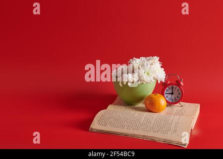 on a red background white flowers in a cup stand on a book with an alarm clock Stock Photo