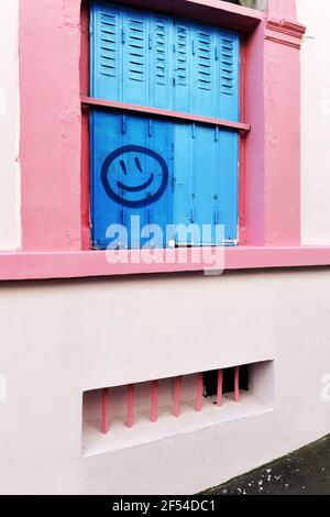 a pink-framed arched window with bright blue shutters and a blue spray-painted smiley face graffiti, located in Montmartre, Paris, France Stock Photo