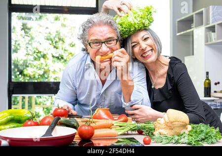 Senior couple having fun in kitchen with healthy food - Retired people cooking meal at home with man and woman preparing lunch with bio vegetables Stock Photo
