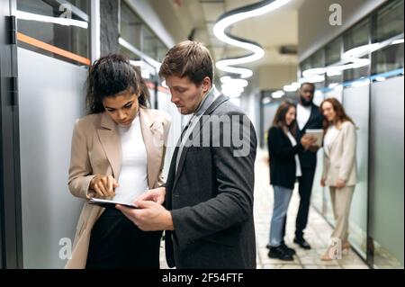 Focused business people are working together in office, using a tablet. Multiracial serious young adult colleagues are developing important project or startup, teamwork concept Stock Photo