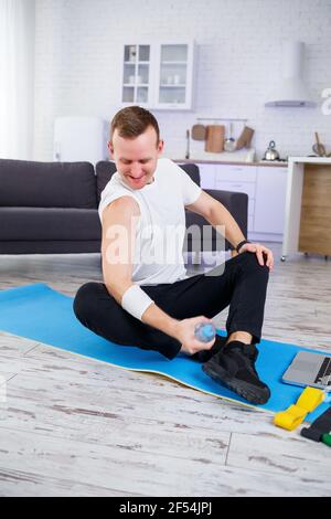 Handsome young man sitting on the floor and holding bottle of water while having online workout Stock Photo