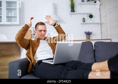 A young successful man is sitting at home on the couch with a laptop and working. Remote work during quarantine. Stock Photo