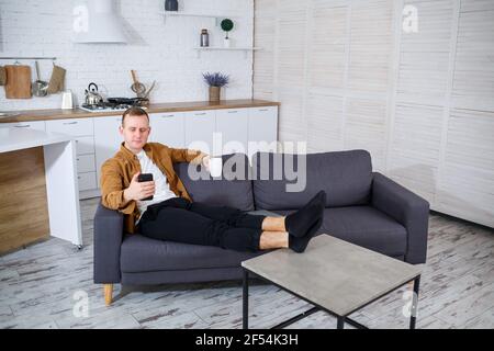 A young successful man is sitting at home on the couch with a phone and talking on a video link. Remote work during quarantine. Stock Photo