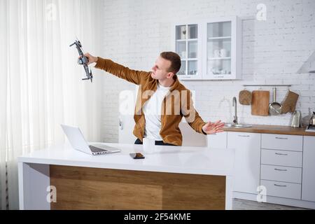 Handsome young businessman working with computer remotely sitting at the table in the kitchen. Nice happy man sitting in the room holding a drone in h Stock Photo