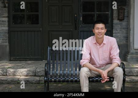 smiling Asian young man sitting on bench outside cafe Stock Photo