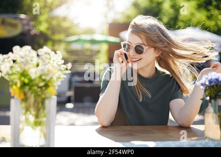 Portrait of young freckled blonde woman talking with her friends on her pink cell phone wearing sunglasses on a sunny day in park. Stock Photo